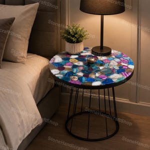 May include: A circular side table featuring a mosaic top composed of vibrant agate stones. The table is supported by a black metal frame and legs. A small potted plant and a lamp are placed on the table. The table is beside a bed.