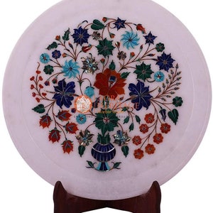 Op de afbeelding: Witte marmeren decoratieve plaat met een bloemeninlegontwerp. Het ingewikkelde patroon bevat rode, blauwe, groene en turquoise bloemen en bladeren. De plaat wordt gepresenteerd op een donkerbruine houten standaard.