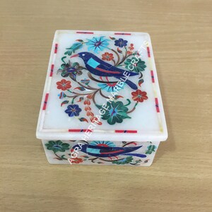 May include: A white rectangular marble box with a lid, adorned with a colorful inlaid design of a blue bird perched on a floral branch. The box features red, blue, and green floral accents. The text "AGRA HERITAGE MARBLE CRAFTS" is visible.