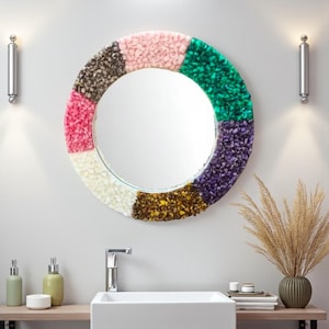 May include: A round mirror with a colorful, textured frame. The frame is divided into segments of pink, green, purple, yellow, white, and brown. The mirror is mounted above a white sink in a bathroom setting.