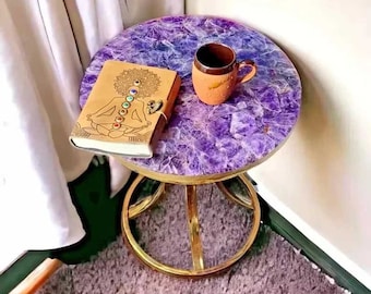 May include: A small, round amethyst table with a gold metal base. A brown leather journal with a mandala design and a brown ceramic mug are on the table.