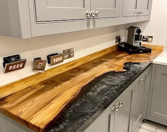 May include: A custom kitchen countertop with a unique wood grain design and a dark, river-like epoxy resin inlay. The countertop is installed with gray cabinets and silver hardware. Electrical outlets are visible on the wall.