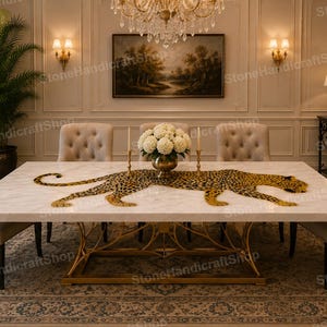 Puede incluir: Una mesa de comedor rectangular de mármol blanco con una base de metal dorado de diseño intrincado similar a ramas. El tablero está decorado con una gran incrustación plana en forma de leopardo dorado y negro y un jarrón de latón central.