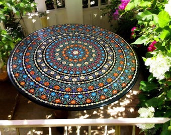 May include: A round table with a black surface and a colorful mosaic design. The mosaic features intricate floral patterns and geometric shapes in shades of blue, green, orange, and white.