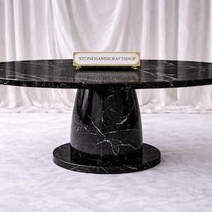 Könnte beinhalten: Ein ovaler Esstisch aus schwarzem Marmor mit weißen Adern. Der Tisch hat einen Sockel und einen runden Sockel. Der Text "STONEHANDICRAFT SHOP" befindet sich auf einem kleinen goldenen Schild auf dem Tisch.