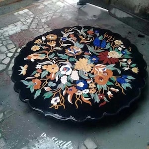 Op de afbeelding: Een ronde zwarte tafelblad met een bloemenmotief gemaakt van ingelegde stenen. Het ontwerp bevat bloemen in tinten rood, oranje, geel, wit, blauw en groen.