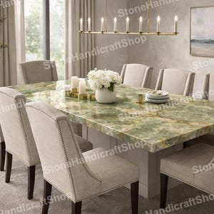 Pode incluir: Uma grande mesa de jantar com tampo de mármore verde, rodeada por dez cadeiras estofadas bege. A mesa está posta com pratos brancos, velas e um vaso de flores brancas. Um candelabro dourado pende por cima.