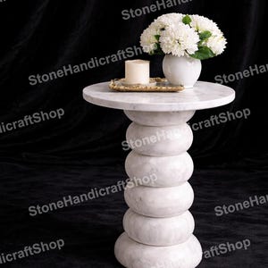 Op de afbeelding: Een witte marmeren bijzettafel met een rond blad en een basis van gestapelde, afgeronde secties. De tafel toont een witte vaas met witte bloemen, een kaars en een gouden dienblad. De achtergrond is zwart.