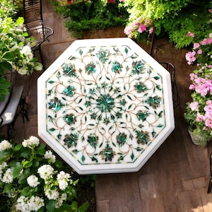 Puede incluir: Mesa de mármol blanco octogonal con un intrincado diseño floral de incrustaciones verdes y doradas. La mesa está rodeada de exuberantes plantas y flores verdes.