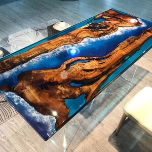 May include: A rectangular coffee table with a distinctive design. The table has a wooden surface with a river of blue resin through the middle. The wood is a natural, warm brown, and the resin is a vibrant blue. The table is supported by a clear glass base.