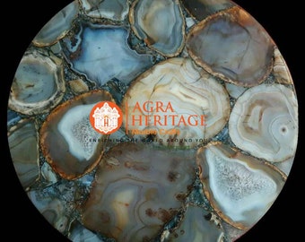 May include: Circular agate stone table top with a mosaic pattern. The stone features a mix of neutral colors, including beige, gray, and white, with brown and gold accents. The table top has the text "AGRA HERITAGE" and "ENRICHING THE WORLD AROUND YOU".