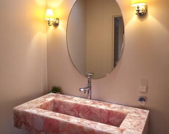 May include: A modern bathroom featuring a rectangular pink stone sink with a chrome faucet, an oval mirror, and two wall sconces. The walls are a neutral color, and the lighting creates a warm ambiance. A small plant adds a touch of greenery.