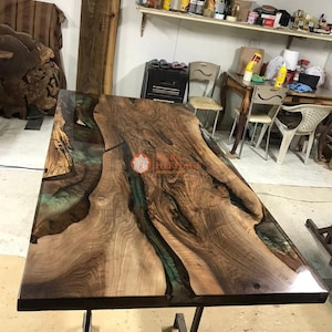 May include: A rectangular live edge wood dining table featuring a polished surface with natural dark brown wood grain and translucent green resin river inlay. The table is supported by a dark metal frame base and displays a central logo.