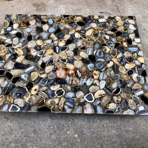 May include: A rectangular slab composed of numerous polished agate slices in shades of black, gray, white, and tan. The stones are arranged in a mosaic pattern with visible grout lines between each piece. A logo is printed in the center of the slab.
