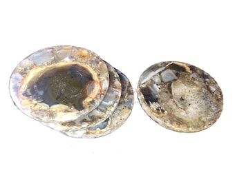 Puede incluir: Posavasos ovalados de piedra natural. Los colores incluyen tonos marrones, grises y crema, con una zona central oscura. La superficie es pulida. Varios posavasos están apilados, uno separado. Cada posavasos es único.