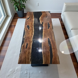 May include: A rectangular coffee table with a dark, glossy black center and natural wood edges. The wood has a warm brown tone with unique grain patterns. The table is supported by a black, rectangular base. The table is approximately 60 inches long.