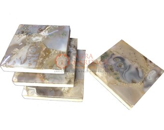 Puede incluir: Un juego de posavasos cuadrados de piedra natural. Los posavasos tienen una superficie pulida con una mezcla de patrones grises, blancos y marrones. Los posavasos están dispuestos en una pila de cuatro, con un posavasos separado.