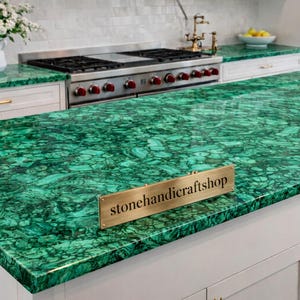 Può includere: Un'isola cucina con un piano di lavoro verde vibrante con un motivo marmorizzato unico. Il piano di lavoro è in pietra lucida, con un cartello in ottone con la scritta "stonehandicraftshop". Mobili bianchi e una stufa in acciaio inossidabile completano il design della cucina.