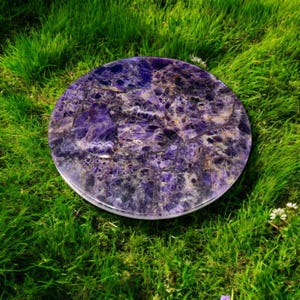 May include: A round amethyst coaster with a purple and white marbled pattern. The coaster is sitting on a bed of green grass.