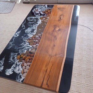 May include: Rectangular coffee table with a unique design. One side features a simulated ocean scene with waves, pebbles, and sand. The other side is a natural wood slab. The table is approximately 48 inches long.