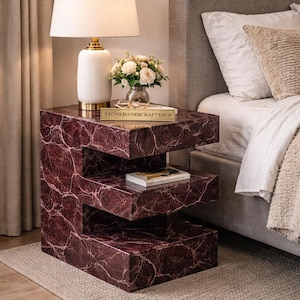 Könnte beinhalten: Moderner Beistelltisch mit einzigartigem, geschichtetem Design. Der Tisch besteht aus einem weinroten Material mit weißen Adern. Eine weiße Lampe mit goldenem Sockel, eine Blumenvase und ein Buch stehen darauf.