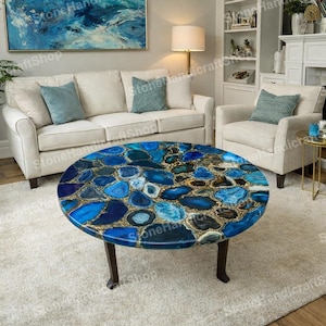 May include: A round coffee table with a mosaic top made of blue and gold-coloured stones. The table has dark brown legs and is set in a living room with a white sofa and chairs. The table is approximately 91 cm in diameter.
