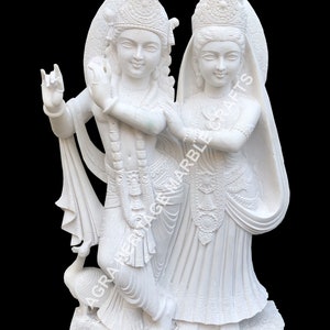 May include: A white marble sculpture of two figures, possibly deities, standing close together. The figures are adorned with detailed clothing and headdresses. A peacock is at the base of the sculpture. The text "AGRA HERITAGE MARBLE CRAFTS" is visible.