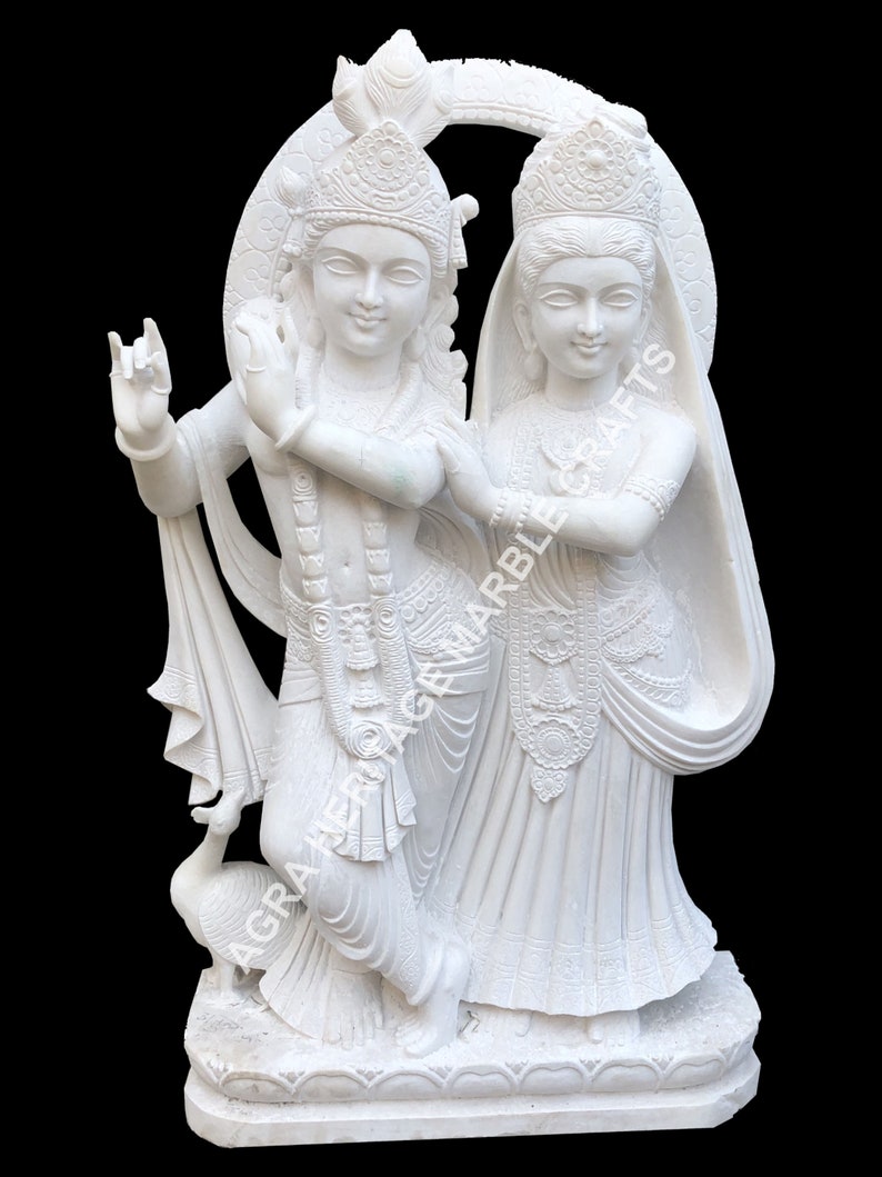 May include: A white marble sculpture of two figures, possibly deities, standing close together. The figures are adorned with detailed clothing and headdresses. A peacock is at the base of the sculpture. The text "AGRA HERITAGE MARBLE CRAFTS" is visible.
