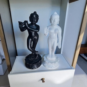 May include: Two statues of religious figures. One is black and playing a flute, the other is white with an open hand. Both are standing on lotus flower bases. The statues are on a white shelf.