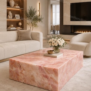 May include: A modern living room featuring a large, rectangular coffee table made of pink marble. The table is the focal point, complemented by a cream-colored sofa, armchair, and a rug. A vase of white flowers and a gold bowl sit on the table.
