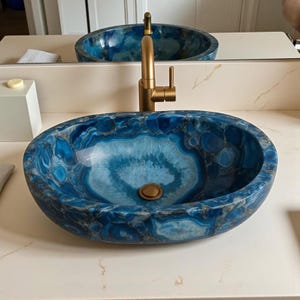May include: Oval-shaped blue and white stone bathroom sink with a gold faucet. The sink has a unique pattern of blue and white swirls and a bronze drain. The faucet is a modern design with a single handle.
