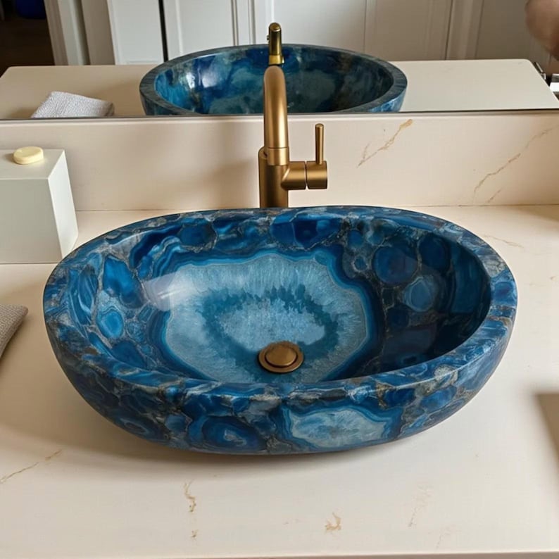 May include: Oval-shaped blue and white stone bathroom sink with a gold faucet. The sink has a unique pattern of blue and white swirls and a bronze drain. The faucet is a modern design with a single handle.