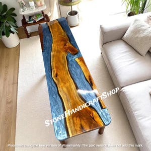 May include: A rectangular coffee table with a wooden center and blue resin. The wood is a warm brown color, and the resin has a translucent, watery appearance. The table has dark brown legs. The text "STONE HANDICRAFTSHOP" is visible.