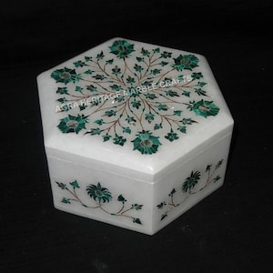 May include: White hexagonal box with a floral design inlaid with green and gold stones. The box is made of marble and has the text "AGRA HERITAGE MARBLE CRAFTS" on the top.