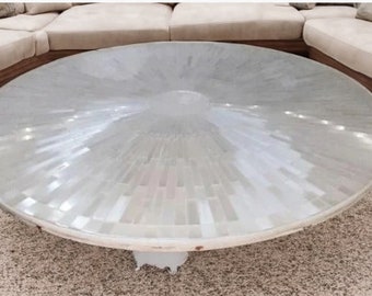 May include: A large, round coffee table with a mosaic-style top. The tabletop is a light, pearlescent color, with a radial pattern. The table has a white base and is surrounded by a beige sectional sofa.