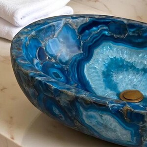 May include: A blue agate stone sink with a polished finish. The sink has a unique, organic shape and a brass drain. White towels are stacked in the background. The sink's interior features concentric patterns of blue and white.