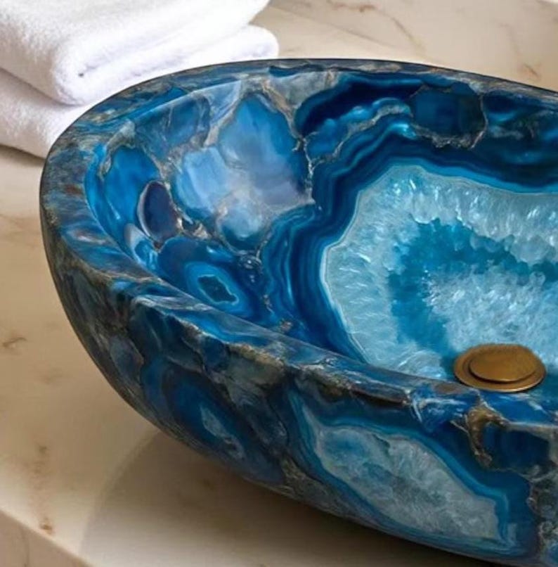May include: A blue agate stone sink with a polished finish. The sink has a unique, organic shape and a brass drain. White towels are stacked in the background. The sink's interior features concentric patterns of blue and white.