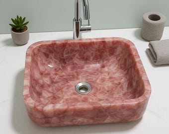 Può includere: Un lavabo da bagno quadrato in pietra color rosa con finitura lucida. Il lavabo presenta un motivo a mosaico e uno scarico circolare. Un rubinetto cromato è montato sopra il lavabo. Una piccola pianta grassa in vaso, un rotolo di carta assorbente e un asciugamano grigio piegato sono nelle vicinanze.