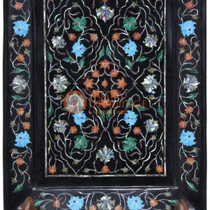 Puede incluir: Bandeja decorativa rectangular negra con incrustaciones florales. La bandeja presenta un detallado patrón floral en tonos azules, naranjas, verdes y plateados sobre un fondo negro. Se apoya en un soporte de madera.