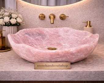 May include: A pink, organically shaped stone sink with a gold drain. The sink is set on a countertop with a gold faucet and fixtures. A sign that reads "stonehandicraftshop" is visible.