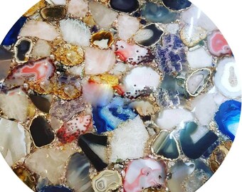 May include: A circular tabletop surface composed of a mosaic of polished agate slices in various shapes and sizes. The stones feature a diverse color palette including shades of blue, pink, white, gray, black, and amber, with gold-toned edges.