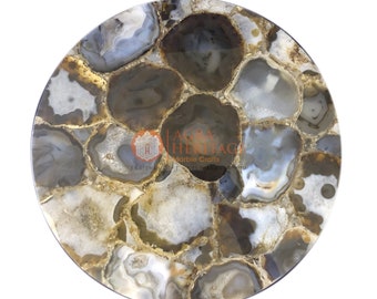 May include: A round table top made of polished agate stone. The surface features a mosaic of gray, white, and brown agate pieces with gold-colored accents. The agate pieces are arranged in a circular pattern, creating a unique and decorative design.