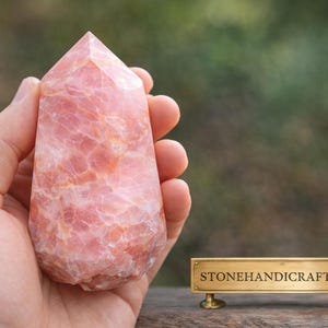 May include: A hand holds a pink, polished crystal point. The stone has a textured surface with variations in color. The crystal is a light pink with darker pink and white marbling. The shop name "STONEHANDICRAFTSHOP" is on a gold sign.