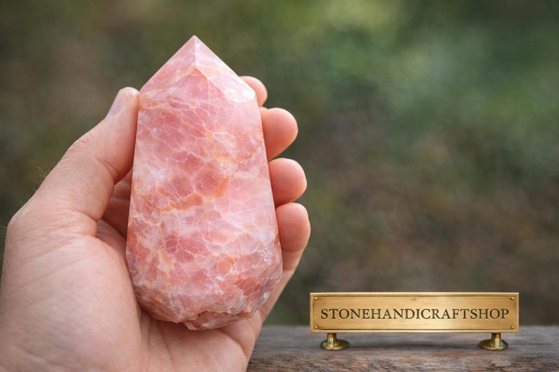 May include: A hand holds a pink, polished crystal point. The stone has a textured surface with variations in color. The crystal is a light pink with darker pink and white marbling. The shop name "STONEHANDICRAFTSHOP" is on a gold sign.