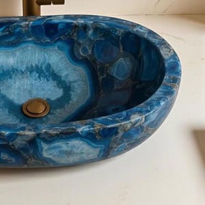 May include: Oval-shaped blue agate stone sink with a brass drain and faucet. The sink's surface features a striking pattern of blue, gray, and white swirls, creating a unique and luxurious bathroom fixture.