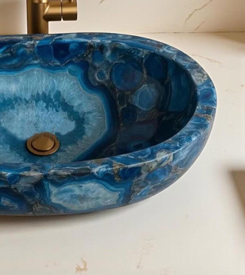 May include: Oval-shaped blue agate stone sink with a brass drain and faucet. The sink's surface features a striking pattern of blue, gray, and white swirls, creating a unique and luxurious bathroom fixture.