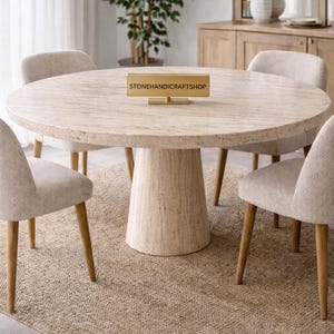 May include: A round dining table with a light-colored stone top and a pedestal base. Four beige upholstered chairs with wooden legs surround the table. The table has a sign that reads STONEHANDICRAFTSHOP.