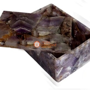May include: A rectangular amethyst box with a lid, showcasing a mosaic of purple, brown, and white crystalline patterns. The box is open, revealing its interior. The box is made of natural stone.
