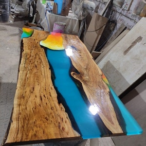 May include: A wooden table top with a blue, orange and yellow epoxy resin river. The resin is polished and glossy. The wood is a light brown colour with dark brown knots. The table top is sitting on a black barrel.