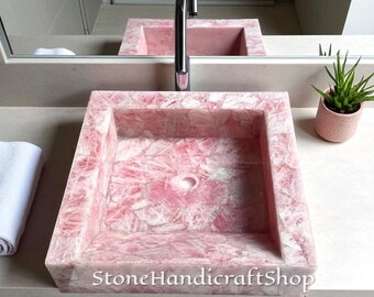 Può includere: Un lavabo quadrato in pietra rosa con una superficie strutturata. Il lavabo ha un bacino rettangolare e uno scarico circolare. È installato un rubinetto cromato. Una piccola pianta in vaso e uno specchio sono sullo sfondo. Il testo "StoneHandicraftShop" è visibile.
